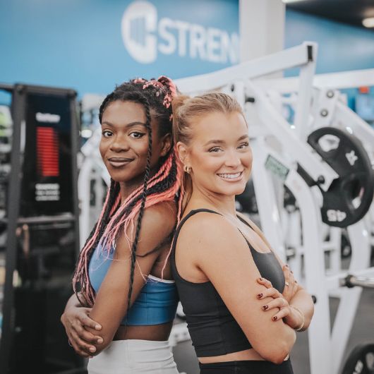 Two women smiling and posing back-to-back at 10GYM Jenks, highlighting gym guest privileges and workout partners.