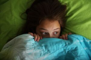 A girl peeks out nervously from under a blue and green blanket, her wide eyes showing fear as she hides in bed.