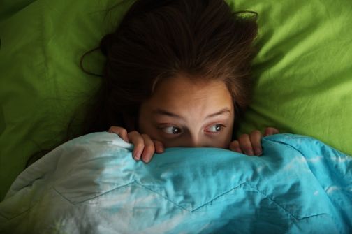 A girl peeks out nervously from under a blue and green blanket, her wide eyes showing fear as she hides in bed.