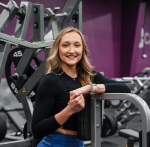 Woman smiling while leaning on a weight machine in 10GYM, wearing a black long sleeve top and blue shorts with strength equipment in the background