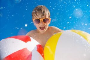 Child wearing sunglasses smiling in water while holding two colorful inflatable beach balls on a sunny day