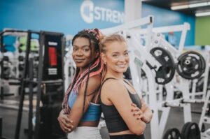 Two women smiling back-to-back in a Freedom Fitness gym beside weight training equipment.