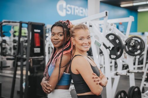 Two women smiling back-to-back in a Freedom Fitness gym beside weight training equipment.