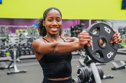 Smiling woman performing a front weight plate raise in a 10GYM location, holding a 10 pound weight plate with both hands. Exercise equipment and free weights are visible in the background.